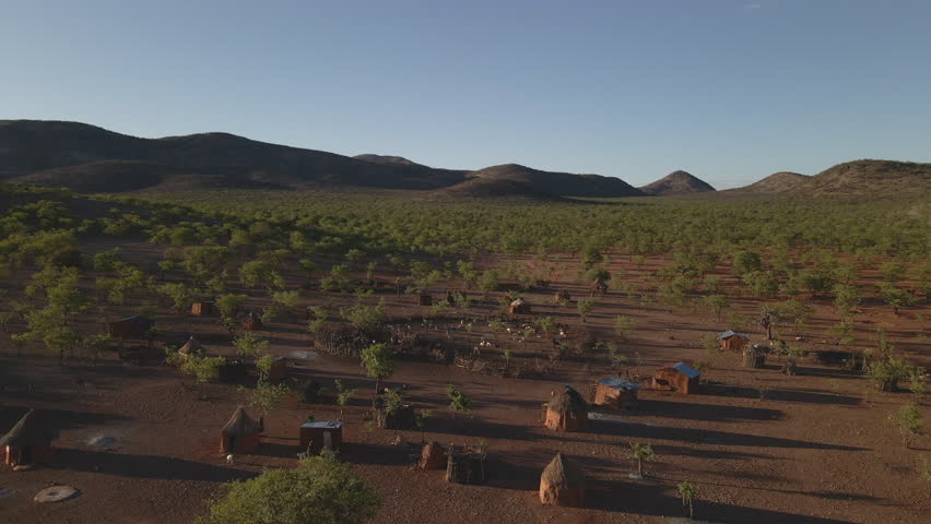 Drone flies over a Himba tribe camp in Namibia during golden hour, revealing red earth, traditional huts, scattered trees, and distant mountains in stunning light.