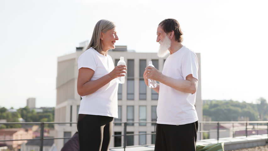 Mature man and woman in casual wear holding bottled water on rooftop after activity. Shows fitness, hydration, togetherness, urban lifestyle, outdoor exercise. Sunny day with scenic city background. - Powered by Shutterstock - Get 15% off with code: PIKWIZARD15