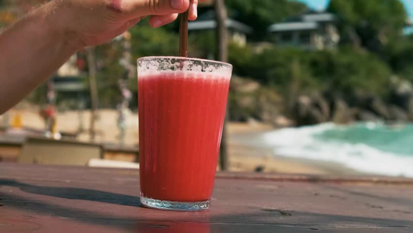 Male hand mixing refreshing red watermelon smoothie in glass with a straw on wooden table in seaside beach cafe, Samui island, Thailand. Cold tropical fruit juice in summer restaurant 