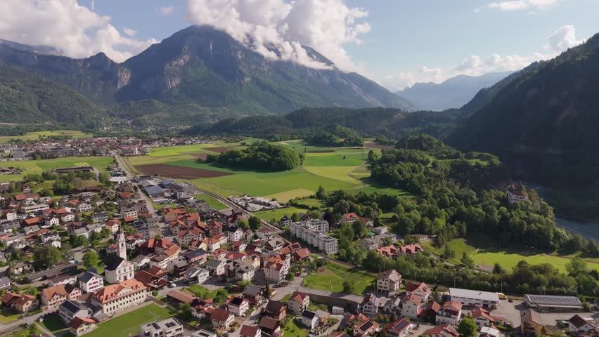 Aerial wide shot of historic houses and Apartments with green farm fields. Rhäzüns Swiss City during sunny day and alps in distance. Beautiful spring day with green trees in Switzerland.