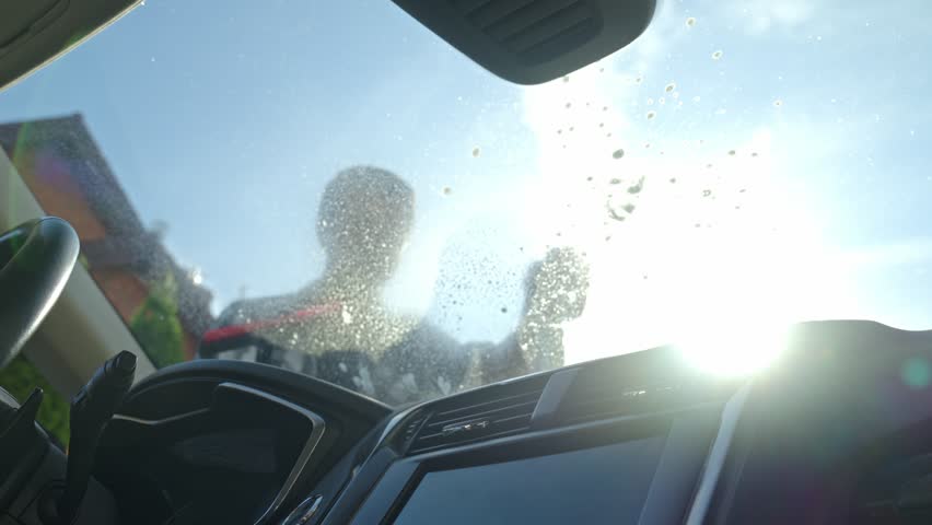 Male washing windshield in sunlight, view from from car inside. Man cleaning car windshield from outside using squeegee. Concept of car care, cleaning services and automotive hygiene