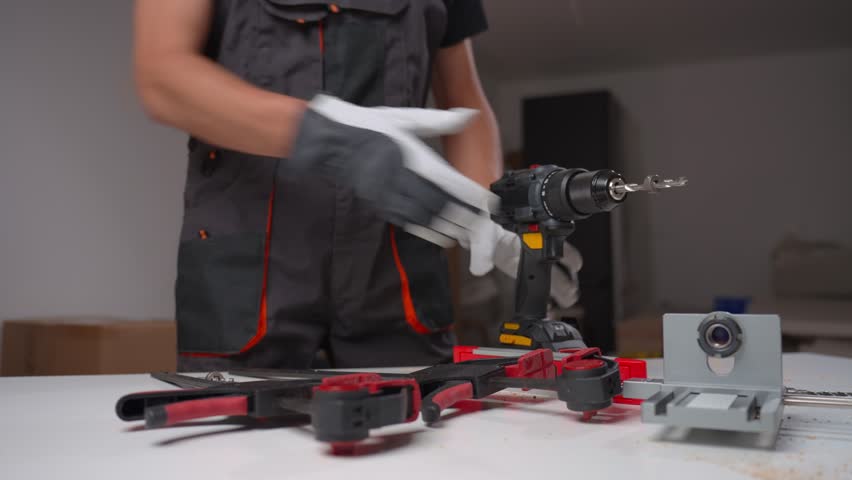 Worker prepares an electric drill for carpentry work in workshop. Man wearing safety gloves and work overalls holding cordless power drill over workbench with tools and components