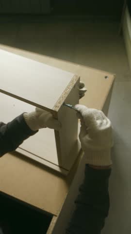 Professional carpenter wearing protective gloves tightening cam lock on particle board furniture using precision allen key during detailed woodworking assembly process