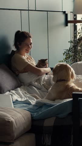 Sunlight filters through the window as a woman serenely enjoys her morning while cuddling her cat, with a dog nearby, in a comfortable and inviting space