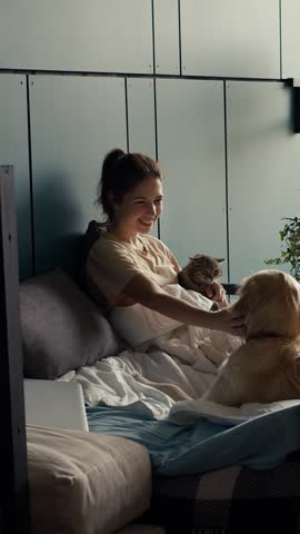 Sunlight filters through the window as a woman serenely enjoys her morning while cuddling her cat, with a dog nearby, in a comfortable and inviting space