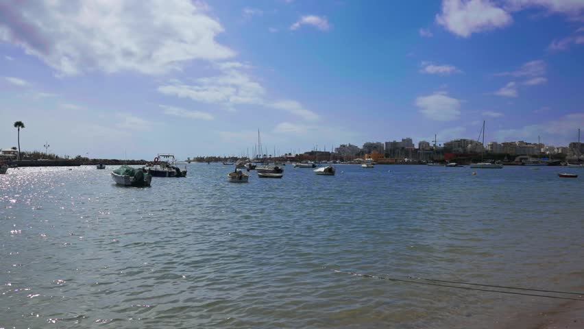 Picturesque marina view showing multiple small boats and vessels anchored in calm waters under clear blue skies with scattered clouds, featuring sunlight reflections and coastal buildings visible in t