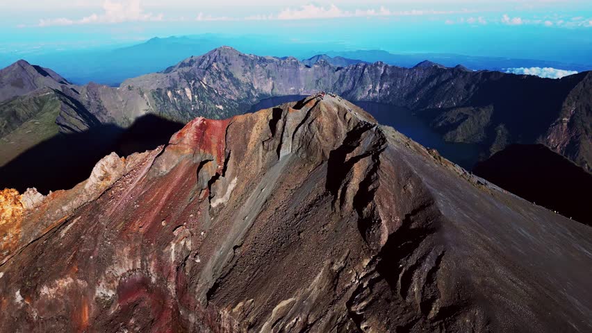 Aerial drone shot of trekkers on Mount Rinjani peak surrounded by volcanic landscape and sparkling crater lake under beautiful natural light.