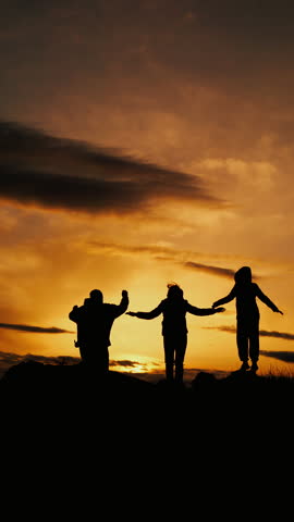 Silhouettes of three tourists against the backdrop of a sunset in the mountains rejoice at their achievement and jump with their hands raised up. Vertical video.