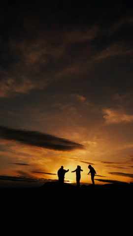 Silhouettes of three tourists against the backdrop of a sunset in the mountains rejoice at their achievement and jump with their hands raised up. Vertical video.