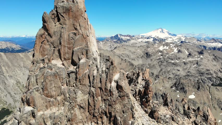 Rocky granite spire of Torre Principal rises sharply with snowy Cerro Tronador in background, drone aerial shot over rugged Andes in Bariloche, Argentina