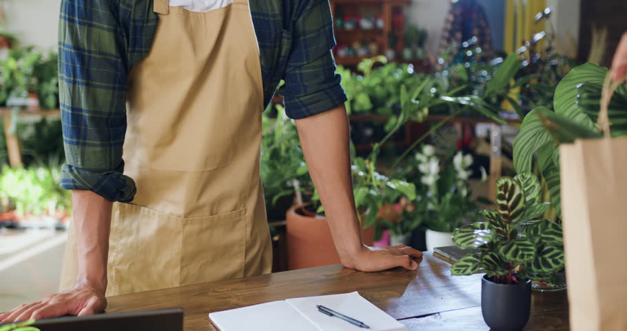 Shopping in flower store. Florist manager working in floral greenhouse. Close up hands man florist worker packs in paper bag and selling house plant in flower shop. People and business concept.
