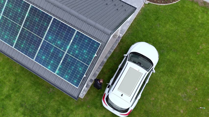 Charging Battery Icon On EV Roof Parked Outside House With Solar Panels - Solar-powered Electric Vehicle Charging. aerial topdown shot