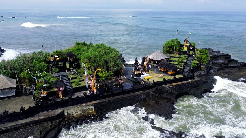 Aerial drone view of Pura Mengening in Cemangi Beach in Bali, showcasing dramatic rock formations, black sand, ocean waves, and a seaside temple.