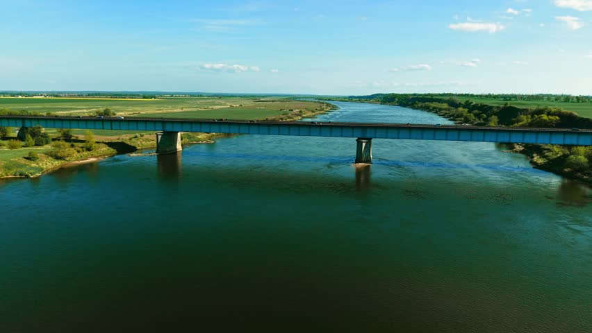 Cars driving across long bridge over wide river in rural landscape. Vehicles moving along elevated road spanning waterway under blue sky. Traffic traveling on viaduct above flowing river in open