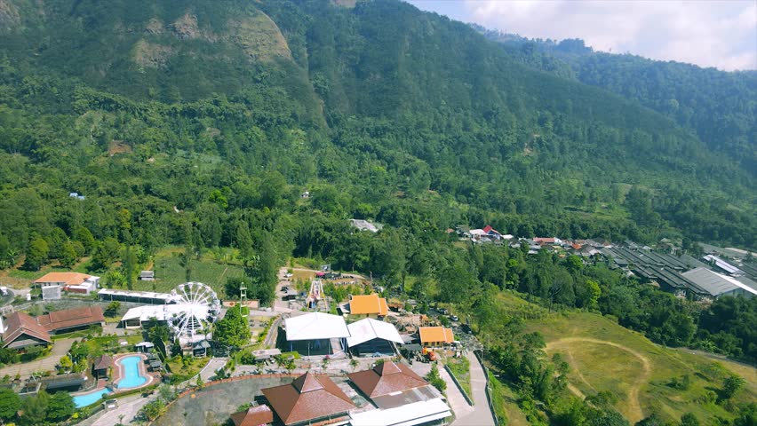 A scenic green valley town is surrounded by mountains and forests under a bright, clear sky.