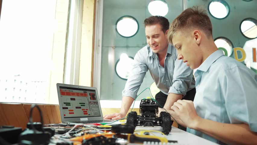 Caucasian teacher pointing at laptop screen and giving advise for programming system or coding prompt while smart high school boy listen and use equipment to repair robot at STEM classroom