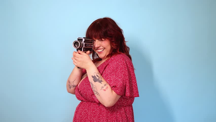 Body positive curvy woman with auburn hair and tattoos wearing a red dress smiles while filming with a vintage 8mm camera, isolated on a blue background