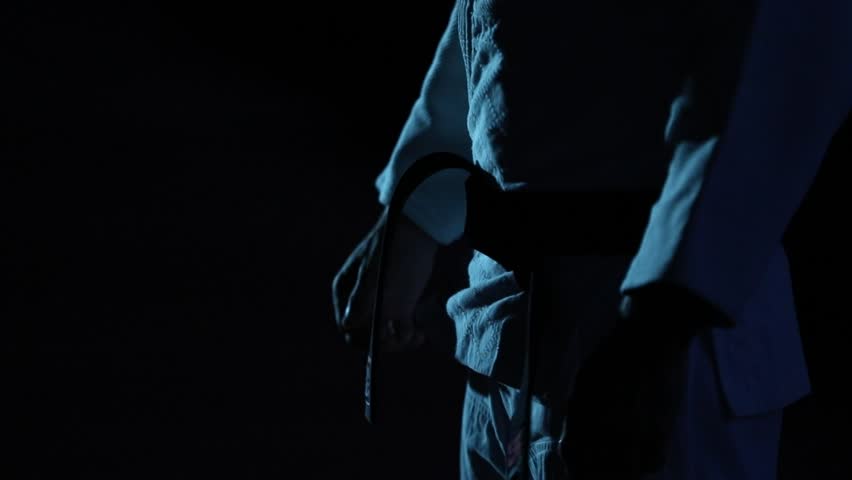 Close-up: Martial artist in meticulously ties their black belt in slow motion. Dark, moody atmosphere with cool blue lighting emphasizes focus and pre-fight ritual.