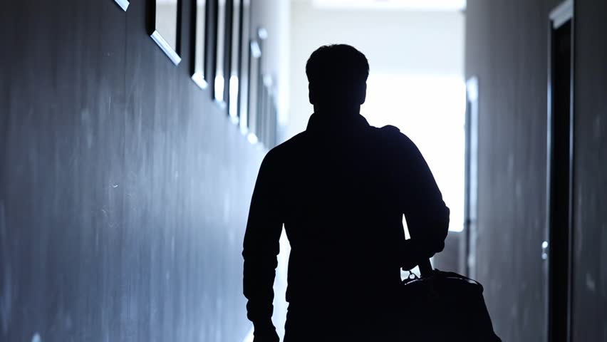 man  walking down dark, blue-lit corridor. He slings a sports bag over his shoulder. Mysterious, atmospheric, purposeful journey.
