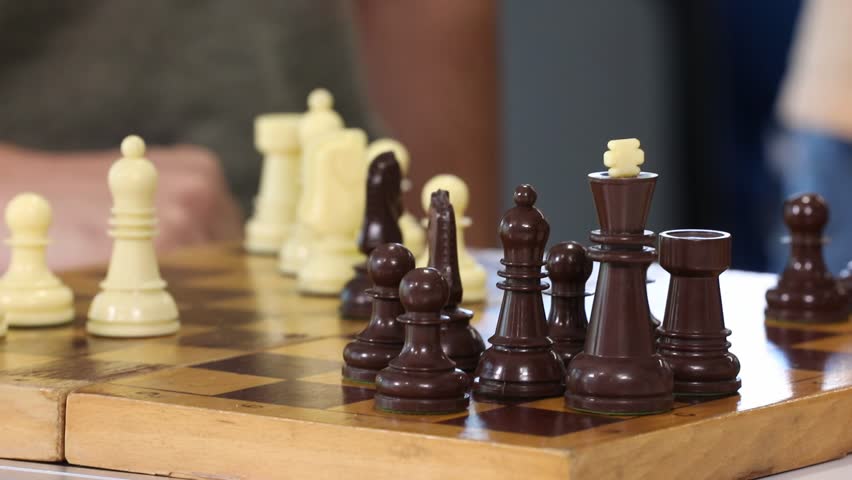 Close-up footage of two players making opening moves in a chess game. Hands strategically position white and black pieces on the wooden chessboard, initiating the intellectual battle.
