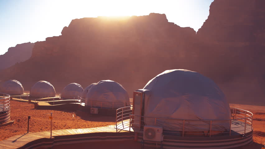 Dome habitats stand on the red desert surface of Mars lit by the sun at sunrise or sunset, representing a human colony.