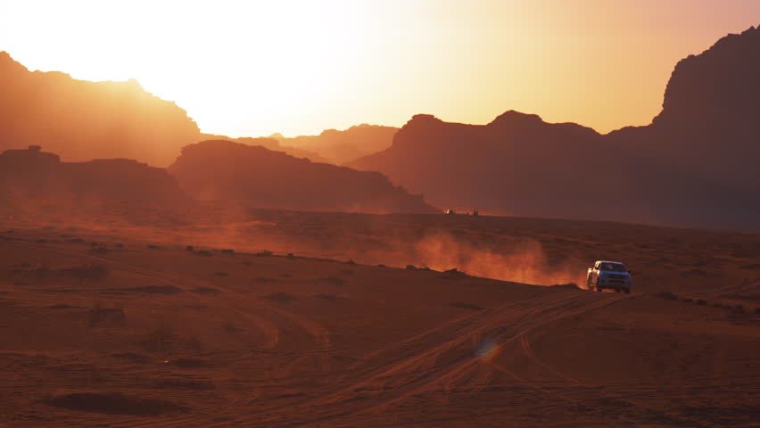Vehicles drive through the red sand desert kicking up dust trails at sunset or sunrise among rock formations in Wadi Rum, Jordan.