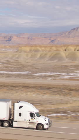 AERIAL: Semi-trailer truck speeds along the motorway running across the desert in Utah. Wintry desert landscape surrounds truck driving down the interstate highway running across rugged US wilderness.