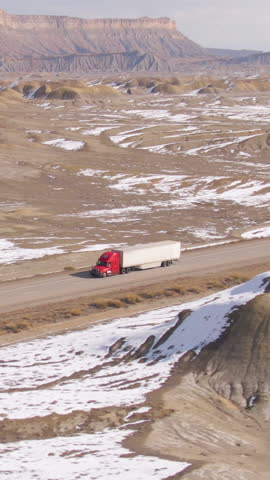 AERIAL: Barren wintry landscape surrounds a cargo truck driving across wintry Utah. Red semi-trailer truck hauls a heavy container across the snowy desert. Truck driving down the interstate freeway.