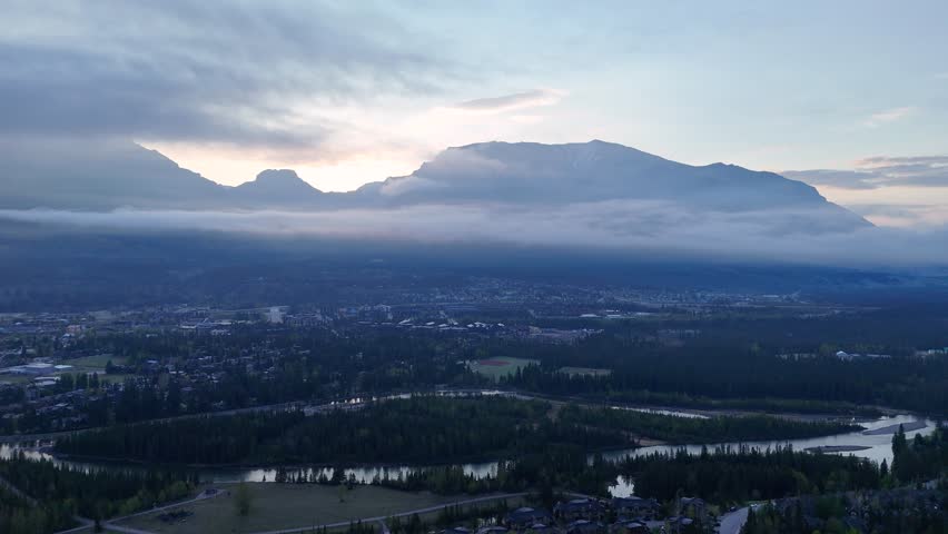 Drone footage of a mountain town at dawn with low clouds wrapping around peaks and a winding river below. Peaceful and cinematic aerial landscape in the Canadian Rockies.

