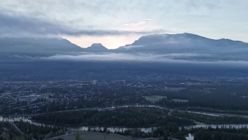 Timelapse aerial footage of a mountain town at sunrise with moving clouds and fog drifting over peaks. River below reflects subtle light changes in this cinematic nature scene.