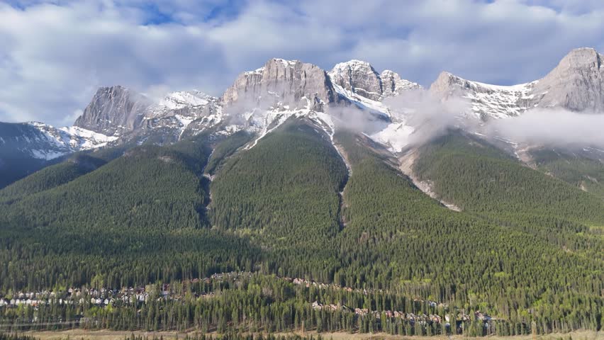 Aerial footage of majestic snow-covered peaks in the Canadian Rockies, with pine forests and clouds drifting across the mountain slopes. Peaceful morning landscape with alpine scenery