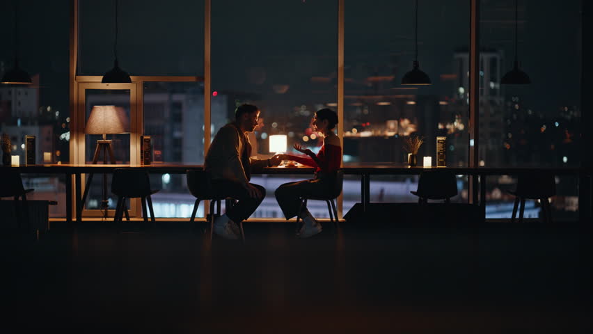 Couple dining in cozy restaurant with night city view. Happy spouses talking at romantic evening date sitting luxury cafe table together. Love pair enjoy relationship spending time in dark cafeteria.