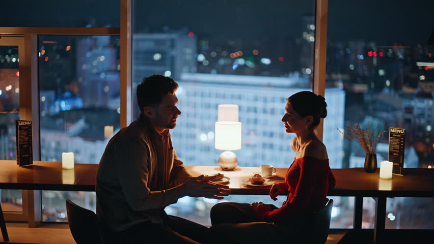 Loving people talking in restaurant with night city view closeup. Bonding happy couple enjoying romantic evening date at cozy cafeteria. Smiling man taking hand beloved woman feeling love tenderness.
