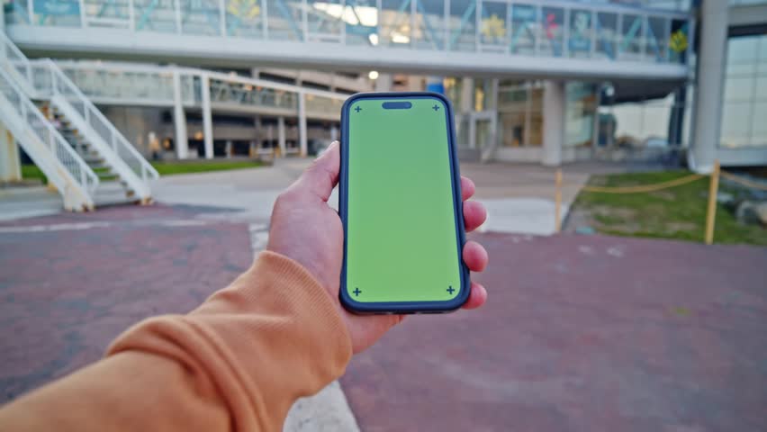 Close-Up POV Of A Man Holding A Smartphone With A Green Screen In Front Of A Sunset Cityscape. The Skyscrapers And Warm Light Add A Modern Touch.
