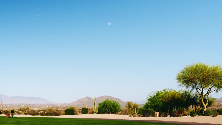 Scenic desert golf course landscape with cacti, mountains, and a pale full moon in the clear blue sky