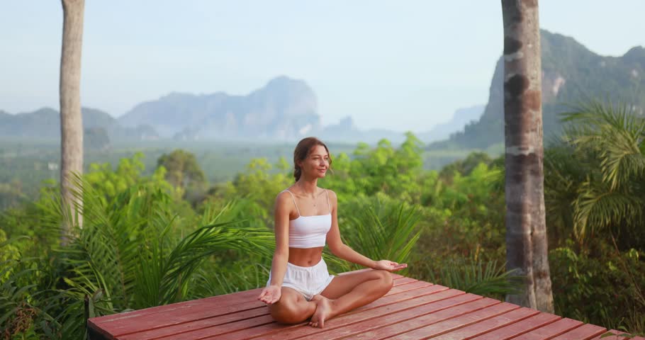 Young Caucasian woman in white outfit doing peaceful yoga poses on a wooden platform in Krabi, Thailand. Ideal for wellness, spa, fitness, travel, and nature lifestyle promotions.