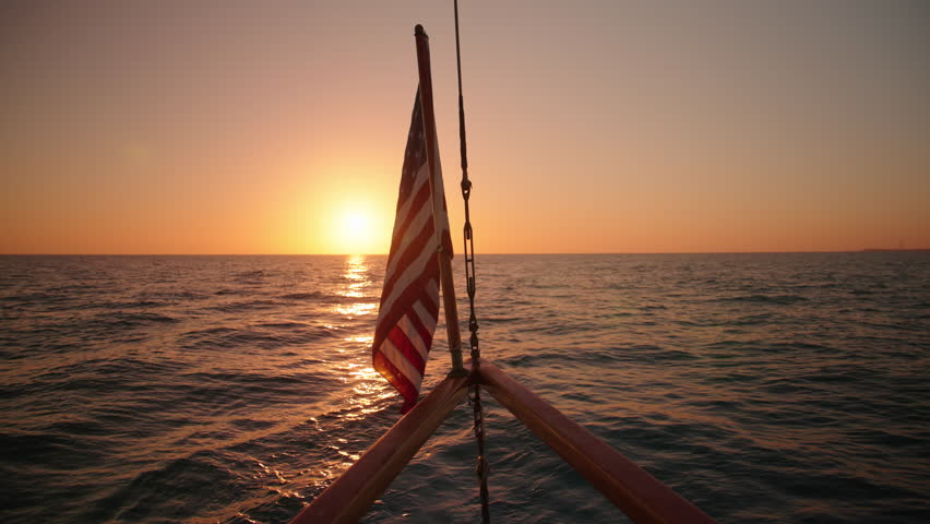 Experience the beauty of an American flag waving on a sailing ship at sunset, captured in slow motion against the stunning backdrop of Key West, Florida, creating a tranquil atmosphere