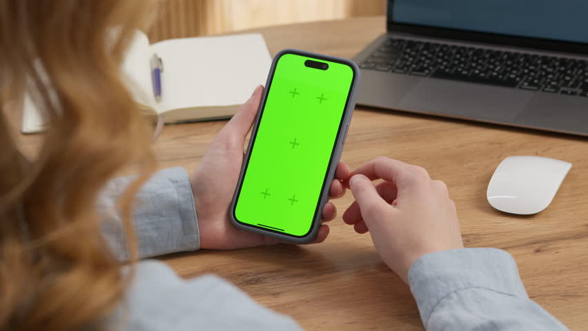 Woman sit use digital tech cell phone, green screen mock up. Hand scroll iphone web site, swipe like photos. Girl hold mobile closeup. Chroma key page. Cozy office background. Online app copy space.