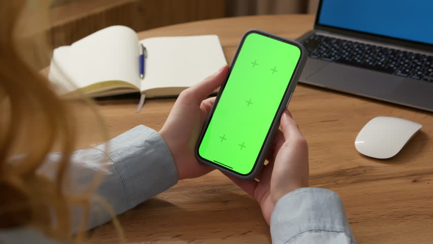 Woman sit use digital tech cell phone, green screen mock up. Hand scroll iphone web site, swipe like photos. Girl hold mobile closeup. Chroma key page. Cozy office background. Online app copy space.
