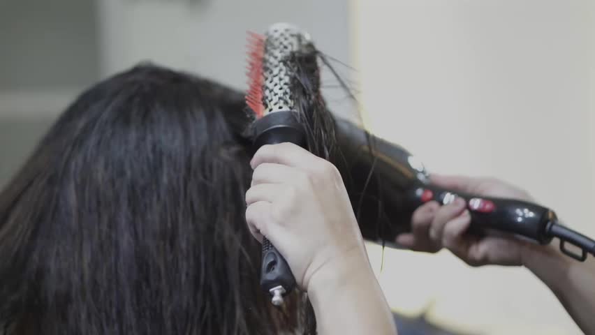A stylist dries a woman's brown hair using a round brush and hair dryer at a salon.