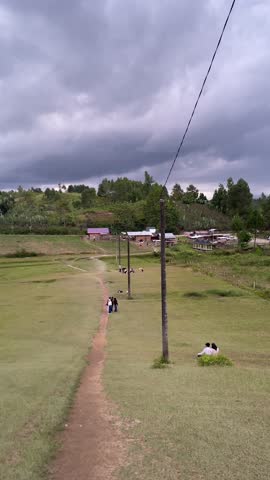 A scenic rural landscape featuring a narrow dirt path lined with electric poles leading to a small village surrounded by hills and trees under a dramatic, cloudy sky. People are seen walking and relax