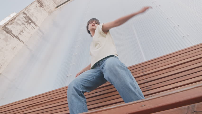 Low angle of short haired young energetic woman looking at camera while hip hop dancing on bench outdoors