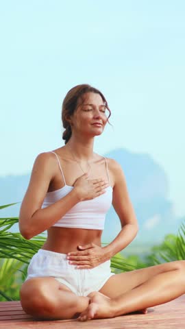 Young caucasian woman in white clothes practicing deep breathing and meditation outdoors with mountains and greenery in the background. Ideal for wellness, spa, and mindfulness campaigns.