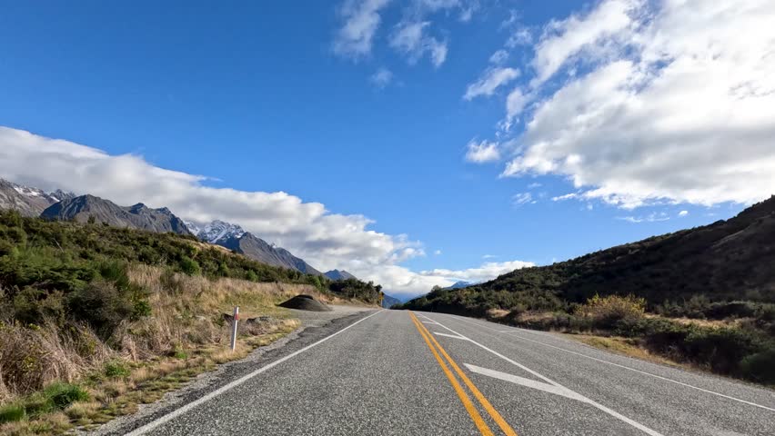 Vehicles travel along a picturesque mountain road in Queenstown, New Zealand, under clear blue skies with scattered clouds