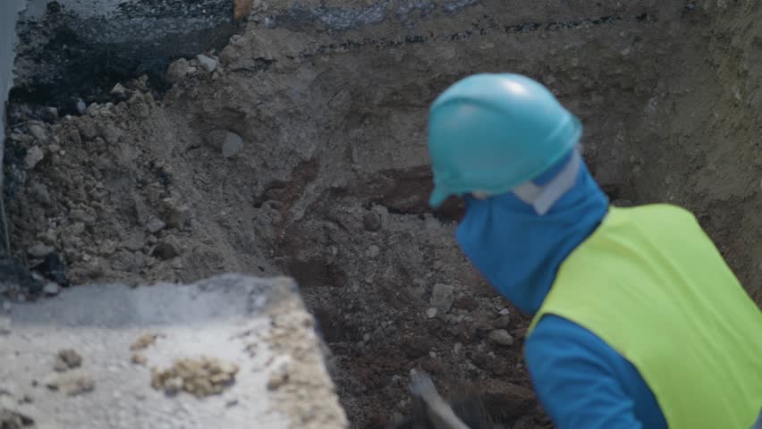 Close-up of construction worker in PPE using shovel to dig trench, hard manual labor from infrastructure or utility worksite.