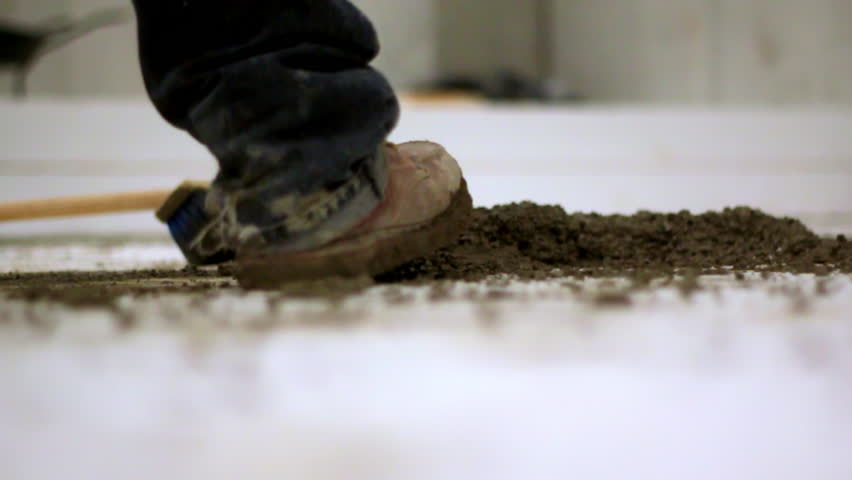 Close-up of a construction worker stepping near a broom and a pile of wet concrete on a white floor, suggesting cleaning or leveling activity during a building process