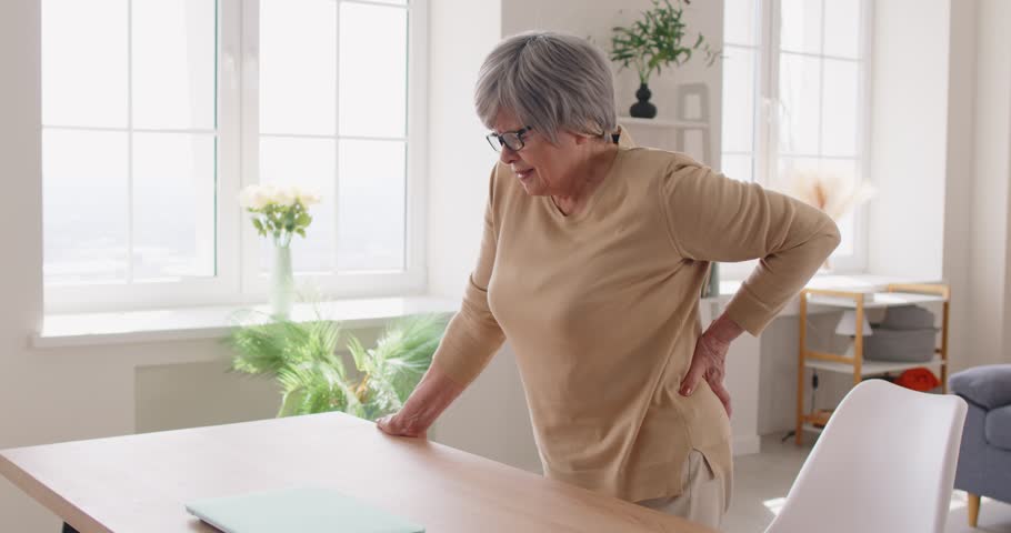 Elderly gray haired woman holding her lower back, suffering from pain. Senior lady leaning on table, experiencing intense discomfort from backaches and lumbar pain, feeling strong spasm in her spine.