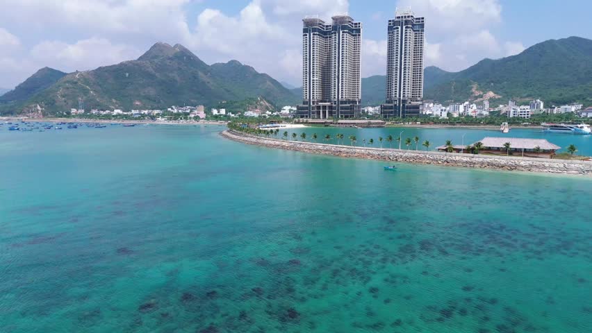 A drone flying over turquoise water towards Royal Marina Center Nha Trang, passing on breakwaters with palms in Nha Trang, Vietnam with mount backdrop