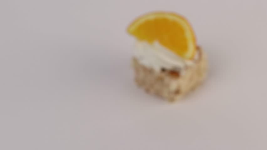 A single mini cake topped with piped cream swirl and a fresh orange slice. The golden-brown sponge contrasts with white frosting. The citrus garnish adds vibrant color accent. The clean white 