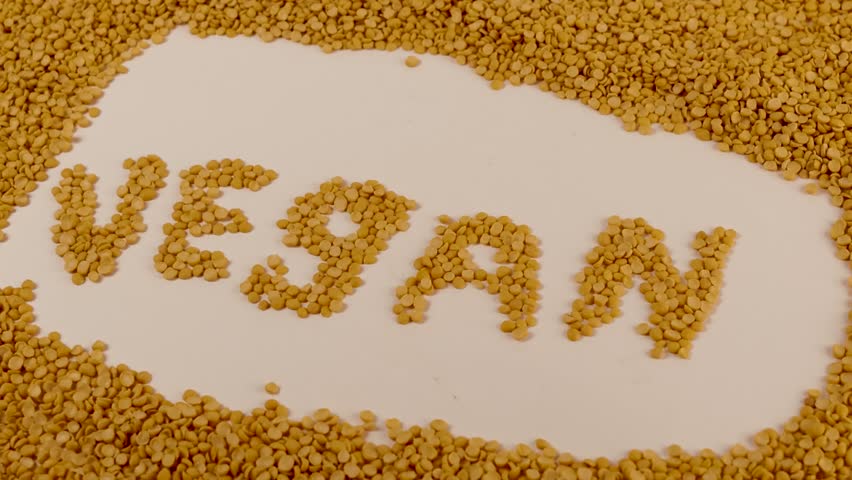 Yellow split peas form the word VEGAN on a seamless white surface. Individual legumes align to create clean letter shapes. Extra peas frame the text in a natural border. Soft overhead lighting softens
